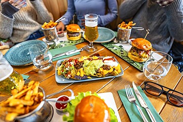 Plate of loaded nachos takes centre stage on a restaurant table, surrounded by burgers, fries, and beers, as friends gather for a casual meal
