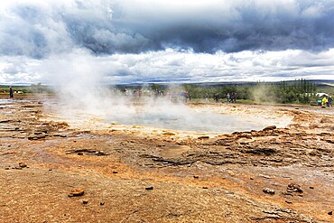 Steaming Strokkur, tourists waiting for eruption, high temperature area Haukadalur, tourist attraction at the Golden Circle, dark rain clouds, Sudurland, Iceland