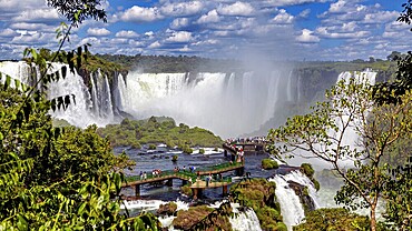 View of a lively waterfall and tourists on a bridge, surrounded by lush greenery and clouds, The Iguazu Falls between Argentina and Brazil