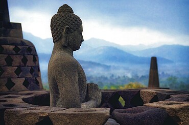 A Buddha statue sits carved in stone with mountains in the background, Candi Borobudur, Borobudur, Java, Indonesia