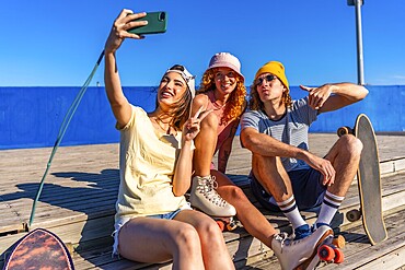 Three young, stylish friends are sitting on a wooden boardwalk, taking a selfie with a smartphone while wearing roller skates and enjoying the sunny weather