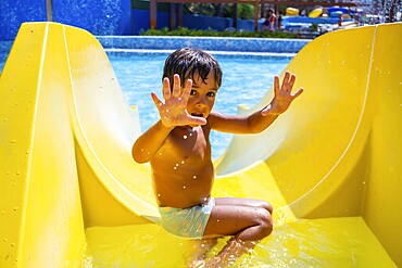 Happy child sliding down yellow water slide in amusement park, having fun during summer vacation