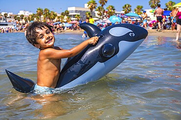 Smiling child having fun riding an inflatable orca toy in the shallow sea water by the beach on a sunny summer day