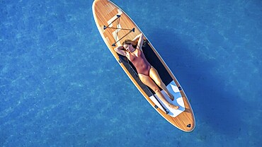 Lying on a paddle board, a woman enjoys the tranquility of the calm blue sea, soaking up the sun and embracing the peaceful summer vibes