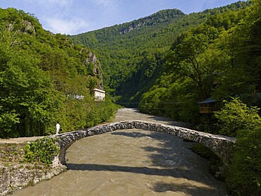 Person crossing a stone bridge over a river, surrounded by green hills and lush forest, aerial view, stone arch bridge Makhuntseti, Adjaristskali River, Adjara Autonomous Republic, Adjara, Adjara, Lesser Caucasus, Georgia