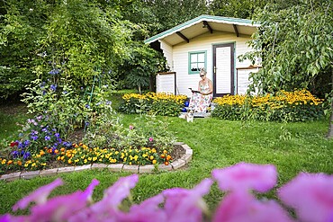 A woman in a dress sits relaxed on the small veranda of a garden shed and reads. Blooming summer flowers in the garden. Germany