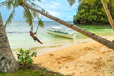 Tourist enjoying a rope swing beneath palm trees on a tropical beach with traditional outrigger boat on kangbangyo island, siargao, philippines