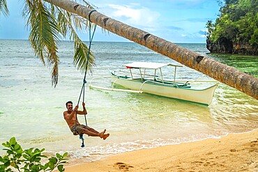 Tourist enjoying a swing on a palm tree with a banca boat on the background, on a beautiful beach in kangbangyo island, siargao, philippines
