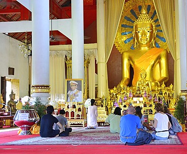 Tourists kneeling and praying in front of a giant golden buddha statue inside a buddhist temple in thailand, demonstrating reverence and cultural immersion