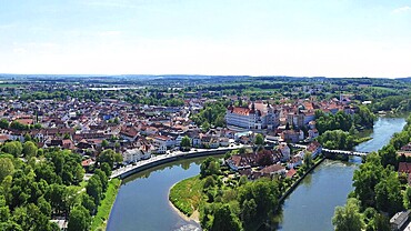 An impressive aerial view of Neuburg an der Donau, a picturesque town in Bavaria. The Danube meanders through the town and forms a small island surrounded by historic buildings with red roofs. Neuburg Castle, which towers majestically over the town and is a historical landmark, is particularly striking. The town is nestled in beautiful natural surroundings with green forests and expansive fields. Neuburg on the Danube, Bavaria, Germany