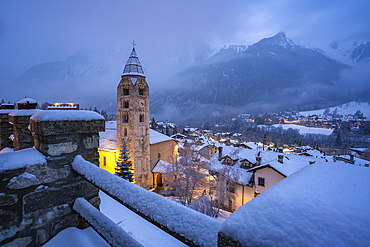 View of Church of Saint Pantalon and the snow covered town centre and mountainous background in winter at dusk, Courmayeur, Aosta Valley, Italy, Europe