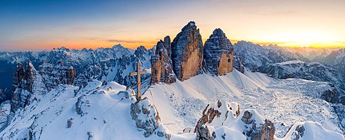 Summit cross on snow capped Monte Paterno with Tre Cime Di Lavaredo on background at sunset, Sesto Dolomites, South Tyrol, Italy