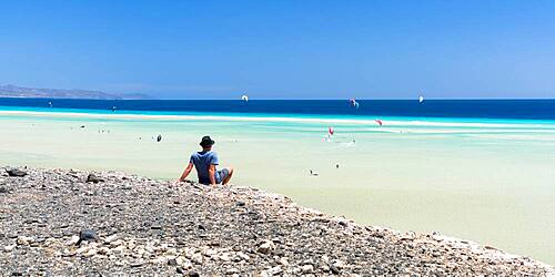 Man sitting on rocks looking at the kitesurfers in the clear sea, Sotavento beach, Jandia, Fuerteventura, Canary Islands, Spain, Atlantic, Europe