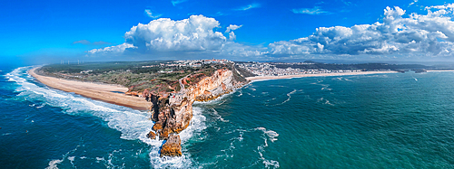 A breathtaking aerial drone panoramic view of Nazare's coastline reveals towering cliffs meeting the ocean, with glorious waves crashing onto the sandy beach, all under a vibrant blue sky