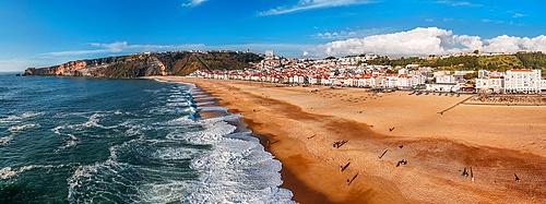 Aerial drone panoramic view of Nazaré coastline showcasing beautiful beach, vibrant surf and rocky pier in Portugal during sunny weather with the iconic rock face on the far left towards North Beach