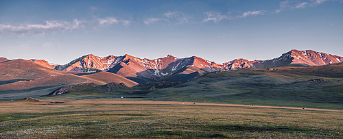 Panorama of a sunset over a snow-capped mountain range in the Song Kol lake area of Kyrgyzstan. The light casts a pink hue across the valley, highlighting the rolling hills and the lush green grass