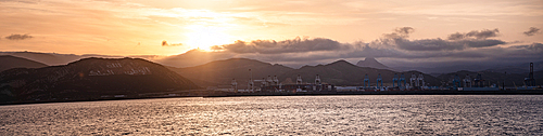 Panoramo of Tangier port at sunrise with industrial cranes and mountains in the background, bathed in soft morning light.