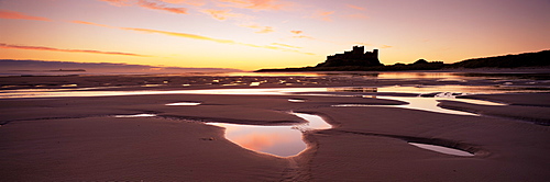 Bamburgh castle in silhouette at sunrise, with rock pools on empty beach, Northumberland, England, United Kingdom, Europe