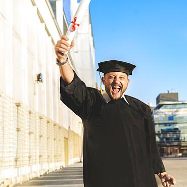 Happy graduated adult man wearing a bachelor gown and a black mortarboard and showing his diploma while looking at camera