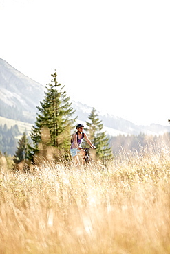 Young woman riding her bike near a meadow on a sunny day, Tannheimer Tal, Tyrol, Austria