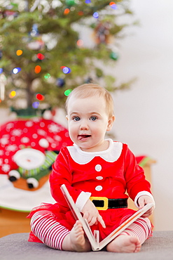 Portrait of a toddler reading a book in her festive Christmas outfit in front of the Christmas tree, Langford, British Columbia, Canada