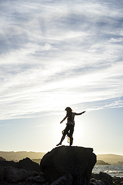 A woman stands balancing on one foot on a rock with a view of the coast at sunset, silhouetted and backlit by the sunlight, San Mateo, California, United States of America