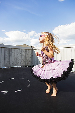 A young girl all dressed up with blond curly hair, sunglasses and jewelry standing on a trampoline in the backyard and showing off, Spruce Grove, Alberta, Canada