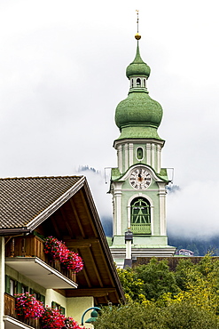 Decorative alpine church clock tower with cloud covered mountains in the background, Dobbiaco, Bolzano, Italy