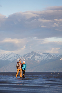 A young couple walking on a beach and holding hands with a mountain range in the distance, Anchorage, Alaska, United States of America