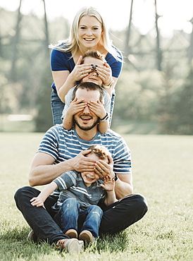 Portrait of a family with young children in a park, standing in a row covering each other's eyes, Edmonton, Alberta, Canada