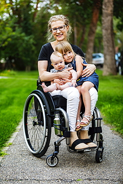 A paraplegic mom carrying her daughters on her lap while using a wheelchair outdoors on a warm summer afternoon and posing for the camera: Edmonton, Alberta, Canada