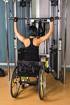 A view from behind of a paraplegic woman working out using a lat pull down machine in a fitness facility; Sherwood Park, Alberta, Canada