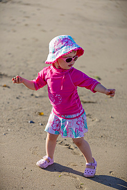 A toddler girl plays at the beach with sunglasses and sun hat, Ambleside Beach; West Vancouver, British Columbia, Canada