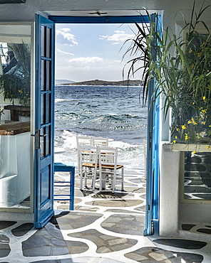 Blue doors wide open to the ocean air and sunlight, with the surf washing onto a patio with chairs, Mykonos, Greece