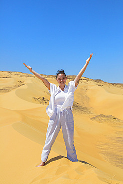 Young woman in white clothing stands with arms raised on the sand dunes of the White Desert Protected Area in Egypt, Egypt