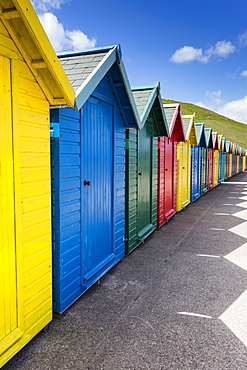 Row of colourful beach huts and their shadows with green hill backdrop, West Cliff Beach, Whitby, North Yorkshire, England, United Kingdom, Europe