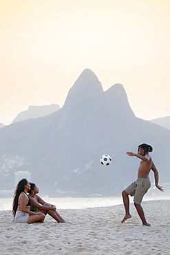 A young black Brazilian playing football with two female friends on Ipanema beach with the Dois Irmaos mountains in the distance, Rio de Janeiro, Brazil, South America