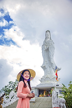 A young Vietnamese woman in a conical hat smiling to camera in front of a statue of Quan Am (Guanyin), Hue, Vietnam, Indochina, Southeast Asia, Asia