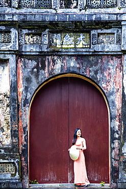 A young Vietnamese woman in a traditional Ao Dai dress and hat standing at the gateway to the Imperial Purple City, Hue, Vietnam, Indochina, Southeast Asia, Asia