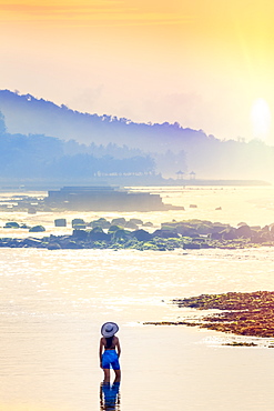 A young woman in a sunhat and shawl standing in a calm ocean as the dawn sun rises, Bali, Indonesia, Southeast Asia, Asia