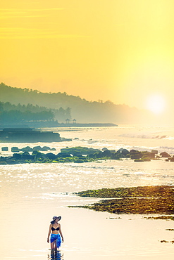 A young woman in a sunhat and shawl standing in a calm ocean as the dawn sun rises, Bali, Indonesia, Southeast Asia, Asia