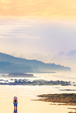 A young woman in a sunhat and shawl standing in a calm ocean as the dawn sun rises, Bali, Indonesia, Southeast Asia, Asia