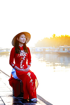 A young Asian woman in a red Ao Dai dress and conical hat smiling and sitting outside next to a river in Hoi An, Vietnam, Indochina, Southeast Asia, Asia