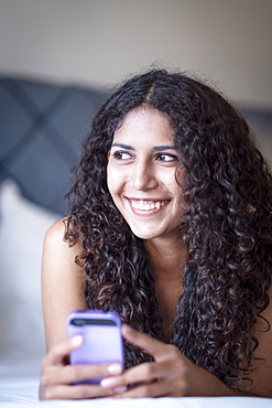 A Latin American woman lying on her bed with her mobile phone and smiling as she looks off camera, Brazil, South America