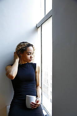 Pensive mixed race woman sitting on windowsill drinking coffee