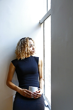 Pensive mixed race woman sitting on windowsill drinking coffee