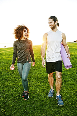 Couple walking in sunny field carrying exercise mat