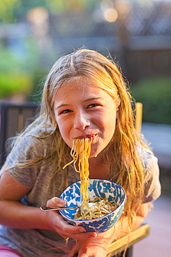 Noodles hanging from mouth of Caucasian girl holding bowl