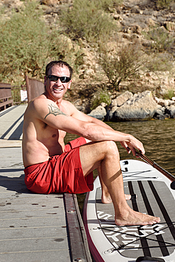 Hispanic man sitting on dock with paddleboard at river