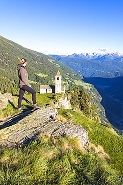 Woman looks at old church perched on mountains, San Romerio Alp, Brusio, Canton of Graubunden, Poschiavo Valley, Switzerland, Europe